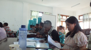 Many people sitting at a dark brown table. There are water bottles, cups, and blue notebooks on the table.