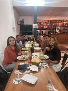 Nine people sitting at a dark brown wooden table eating a meal together. There are plates of food in front of each person and in the center of the table,