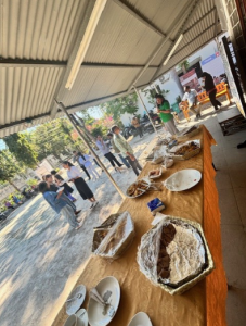 A table full of food dishes with people meeting in the background on a sunny day outside.