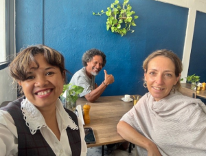 Three people sitting together at a brown table. The wall behind them is blue, and there is a green botanical ornament hanging on the wall.
