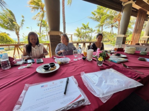 Four people sitting at a table with a bright red table cloth. There are snacks, drinks, and notes on the table.