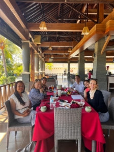 Seven people sitting at a table with a bright red table cloth meeting. There are drinks on the table.