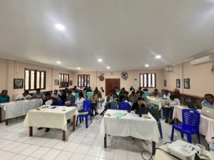 Workshop rooms with nine wooden tables covered in white table cloths. There are two to three people at each table.
