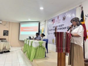 On the right side of the photo, an asian woman with a white collared t-shirt and tan pants is speaking at a wooden podium. On the left side, two people sit at a table with a green and white table cloth